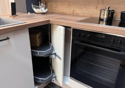 Geöffneter Eckschrank mit schwenkbaren Auszügen neben schwarzem Einbaubackofen und Kochfeld in der Häcker Concept Laser Küche mit Arbeitsplatte in Holzoptik.