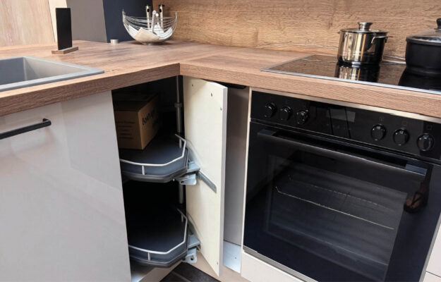 Geöffneter Eckschrank mit schwenkbaren Auszügen neben schwarzem Einbaubackofen und Kochfeld in der Häcker Concept Laser Küche mit Arbeitsplatte in Holzoptik.
